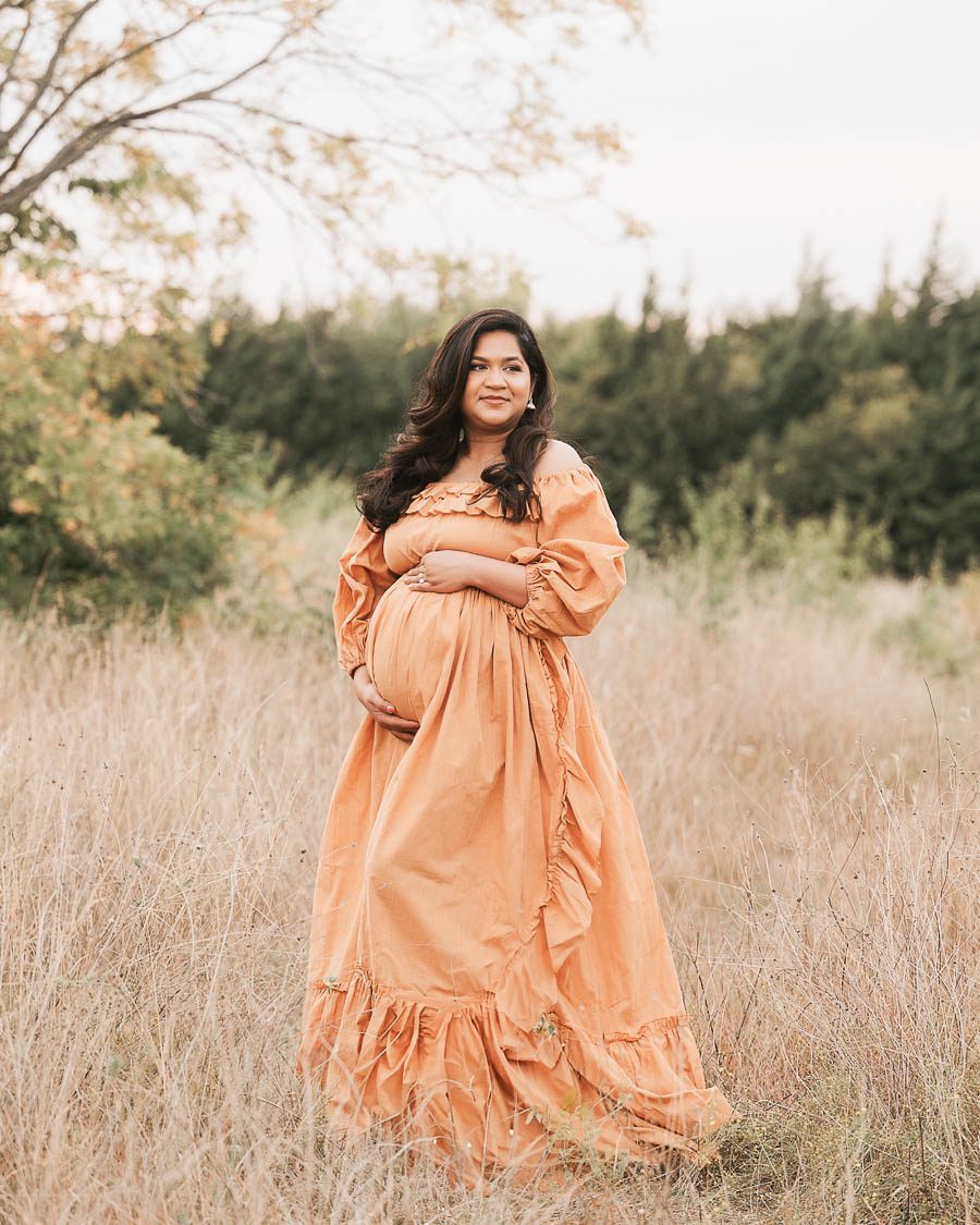 Expecting Mother Wearing Flowing Neutral Gown During An Outdoor Maternity Portrait Session In Nature.