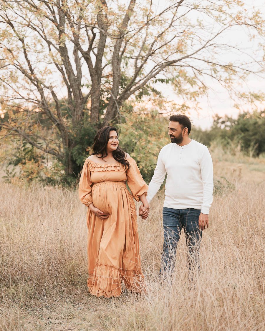 Expecting Couple Walking Together During An Outdoor Maternity Session In A Soft Golden Field.