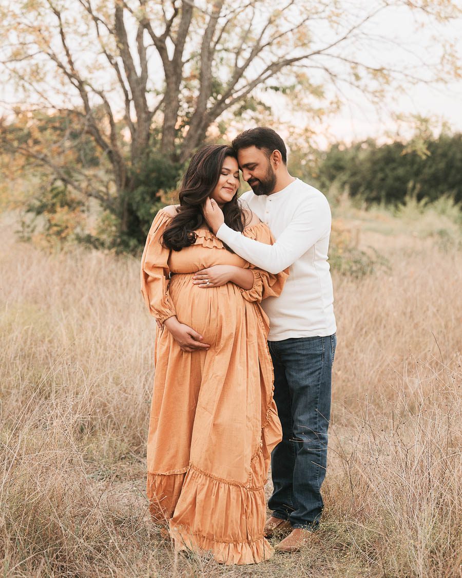 Expecting Couple Embracing During An Outdoor Maternity Session Surrounded By Soft Natural Scenery.