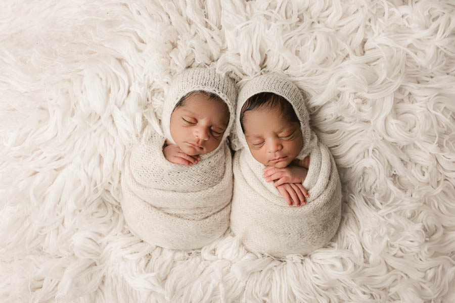 Twin Babies Wrapped In Soft Ivory Fabric Resting Together On Textured Neutral Layers.