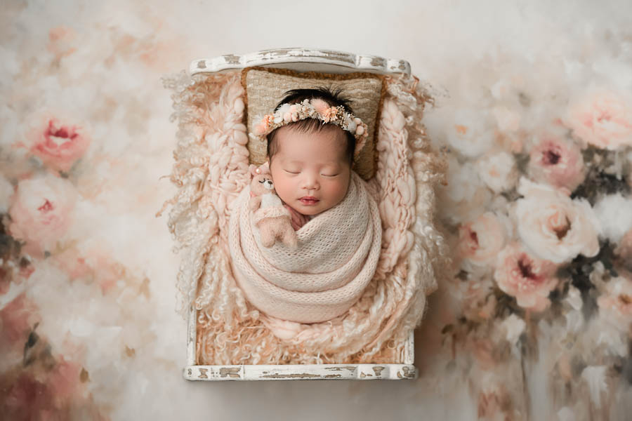 Newborn Baby Sleeping Peacefully In A Styled Newborn Bed Surrounded By Soft Blush Floral Textures.