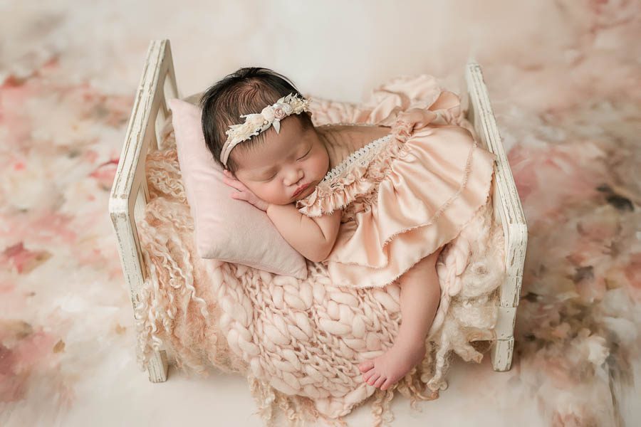 Sleeping Newborn Baby Posed Peacefully In A Delicate Newborn Bed With Soft Neutral Fabrics And Floral Textures.
