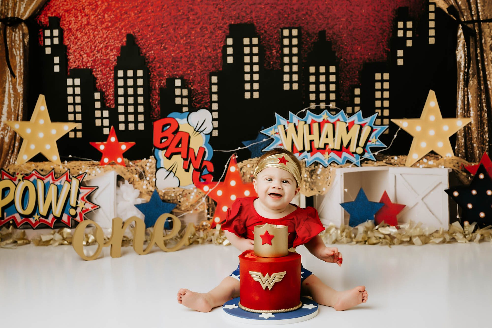 Baby girl in Wonder Woman outfit during superhero cake smash session with themed red and gold cake in Dallas