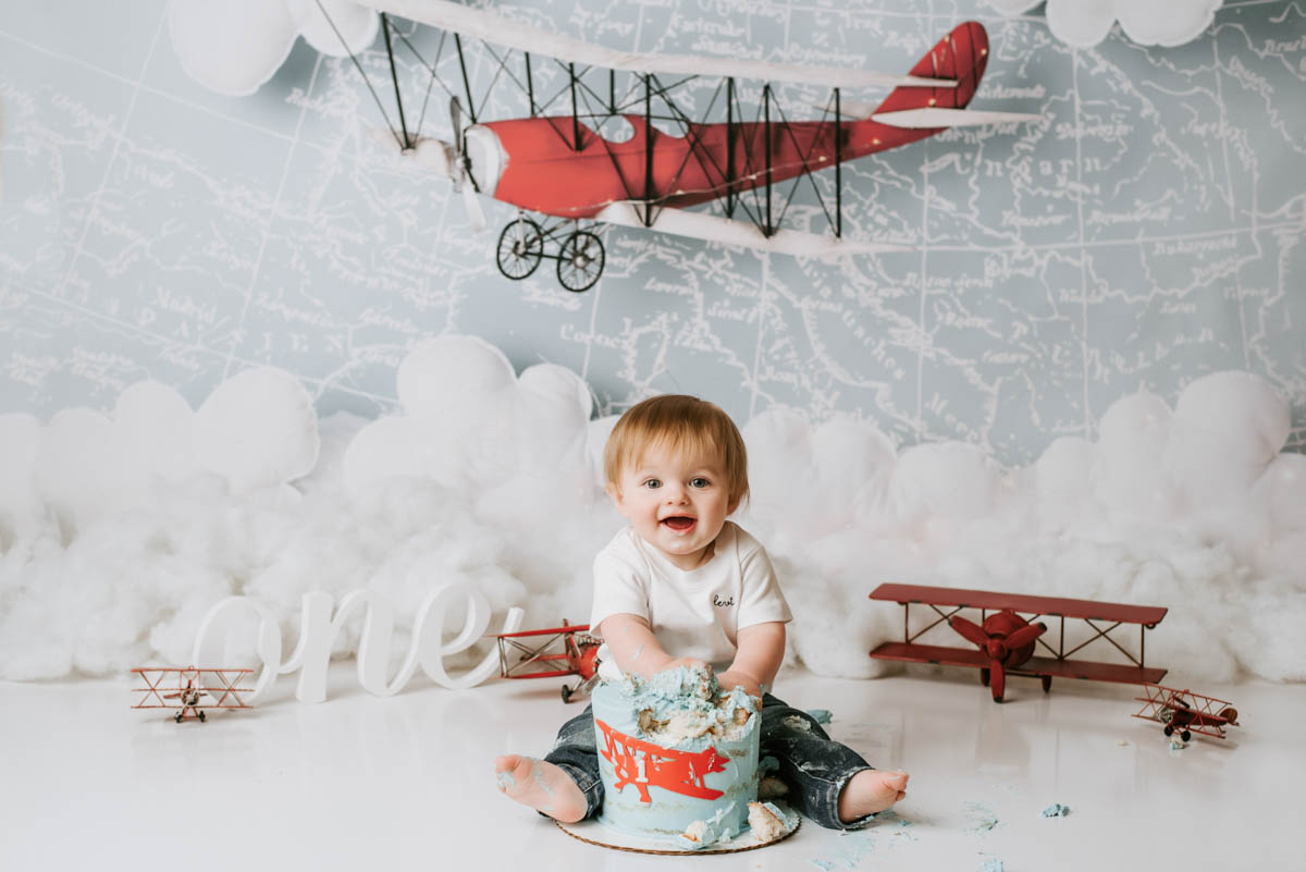 Cake smash photographer in Dallas Lily Hayes Photography capturing a joyful first birthday cake smash with an airplane-themed setup and smiling baby in a professional studio
