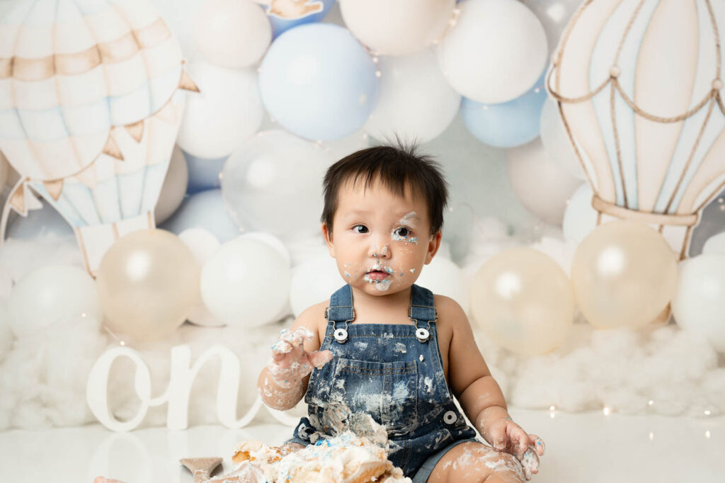 Baby Wearing Denim Overalls During First Birthday Portraits With A Hot Air Balloon Cake Smash Theme
