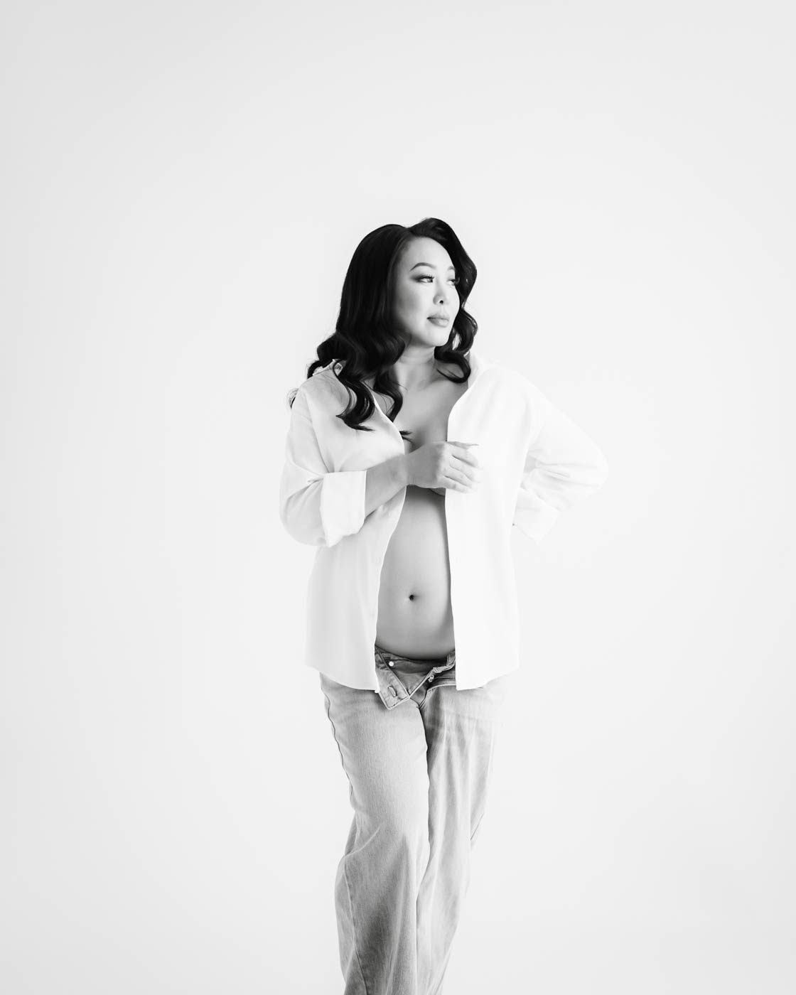 Expectant mother wearing an oversized white shirt and jeans during a minimalist studio maternity photography session in Dallas.