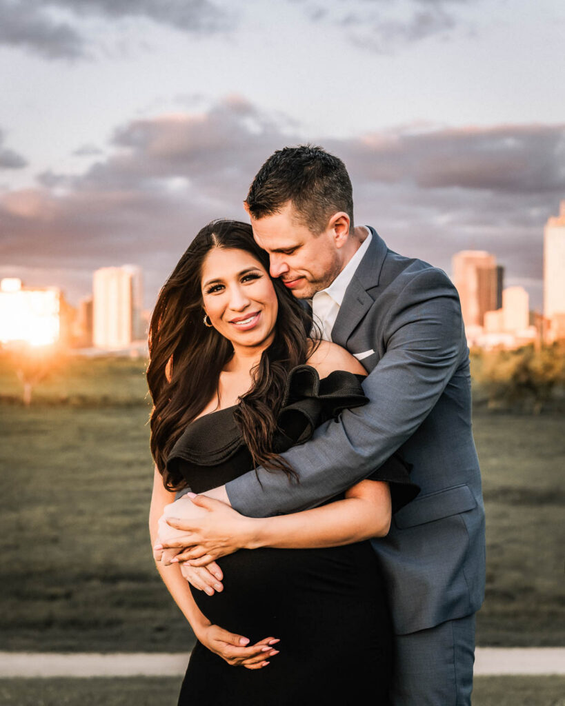 Should Your Partner Be Included In Your Maternity Photos? 4 Dallas Maternity Photographer Portrait Of Expecting Mother In A Black Dress Embraced By Her Partner With The City Skyline Behind Them