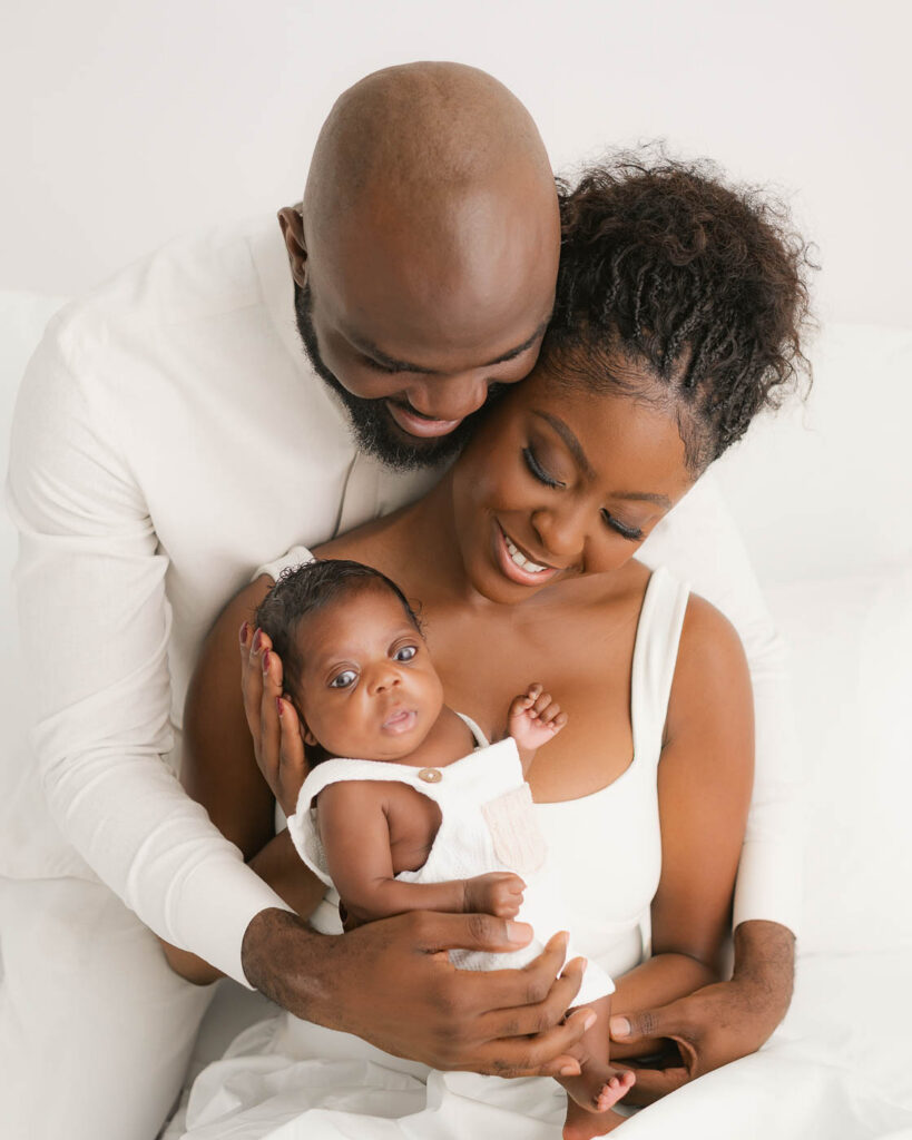 Parents Sitting Together And Holding Their Newborn During A Family Newborn Photoshoot In Dallas