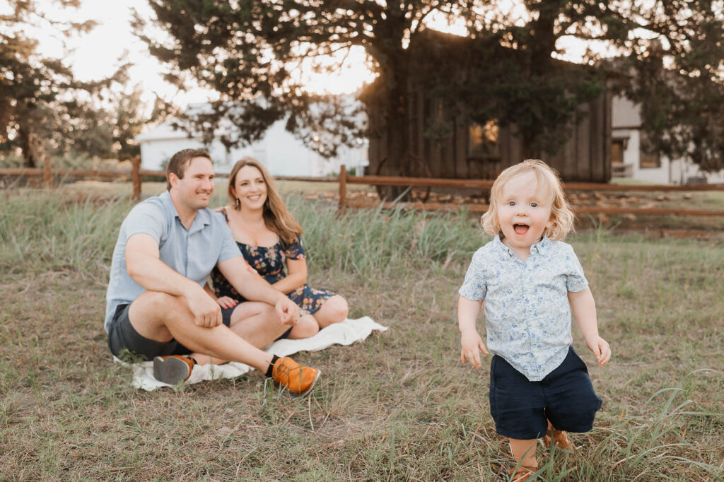 Maternity Photos With A Toddler: What Actually Works 9 Toddler Walking Toward The Camera While His Expecting Parents Sit Behind Him During An Outdoor Dallas Maternity Photography Session.