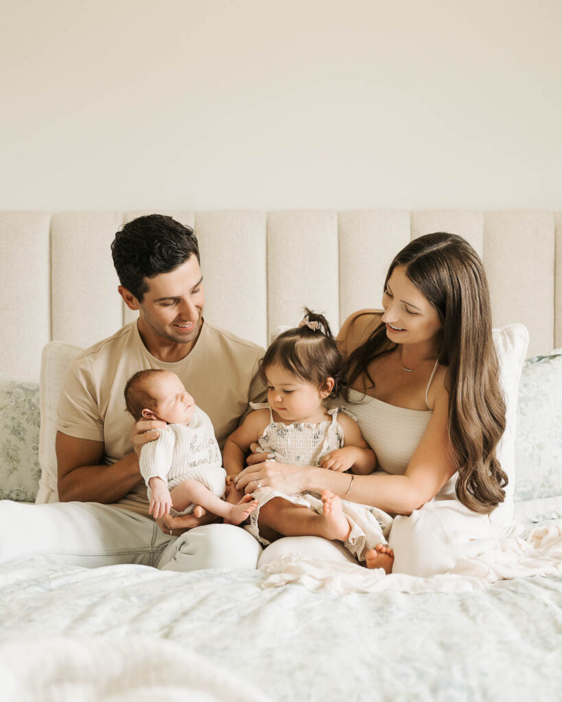Parents Sitting On A Softly Styled White Studio Bed With Their Newborn Son And Toddler Daughter During A Dallas Newborn Family Photoshoot