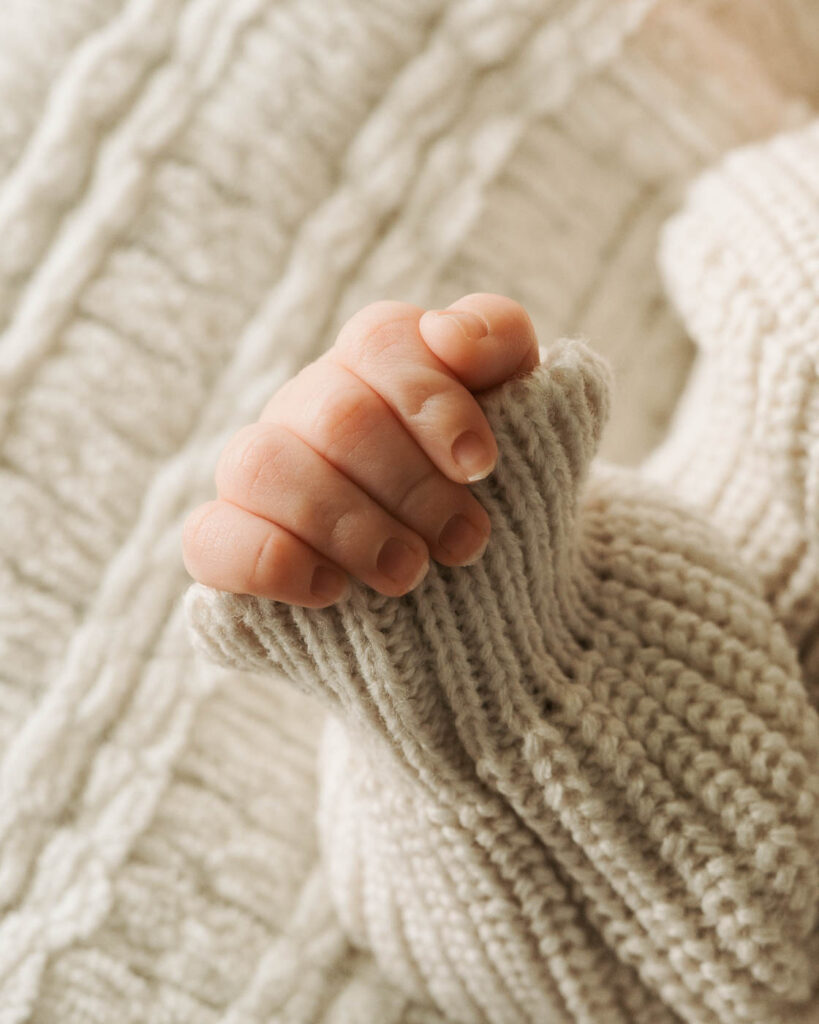 Close-Up Of A Newborn Baby'S Tiny Hand Resting Against The Sleeve Of A Cream Knit Outfit On A Soft Textured Blanket