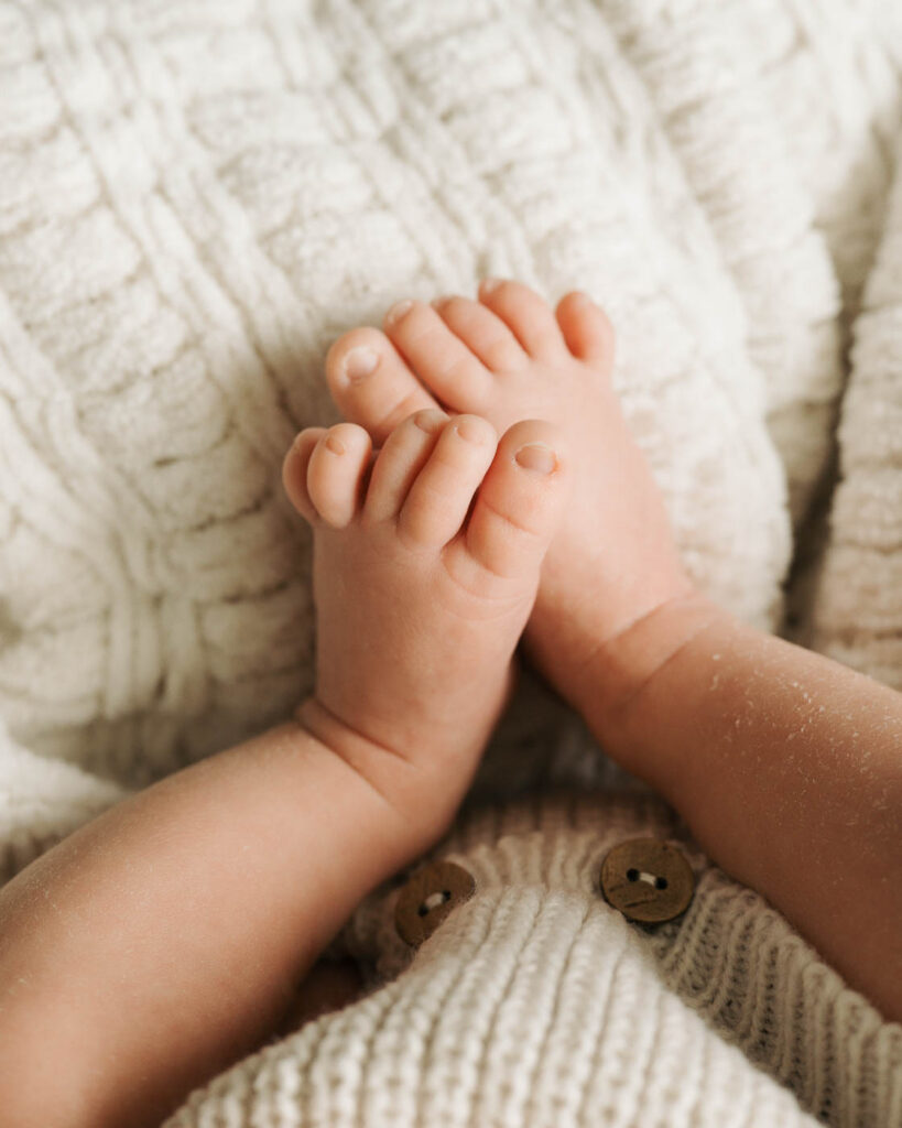 Close-Up Newborn Photograph Of Tiny Baby Feet Gently Held Together Against Soft Ivory Knit Bedding