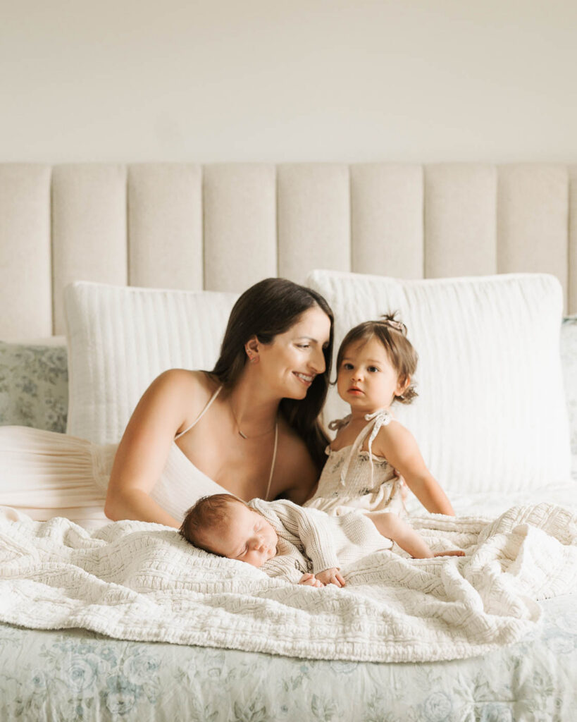 Mother Sitting On A White Studio Bed With Her Toddler Daughter Beside Her And Sleeping Newborn Baby Boy Resting On The Bedding