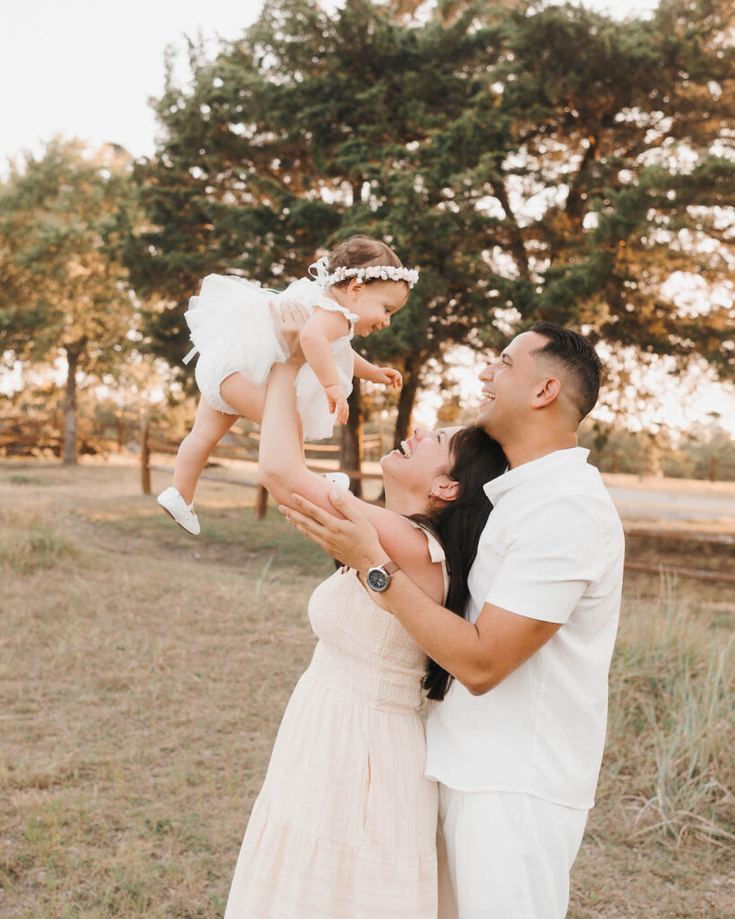 How I Create Natural Family Photos Without Awkward Posing 1 Dallas Outdoor Family Photography Featuring A Mother Lifting Her Toddler Daughter Into The Air While Dad Smiles Beside Them In A Sunlit Field.