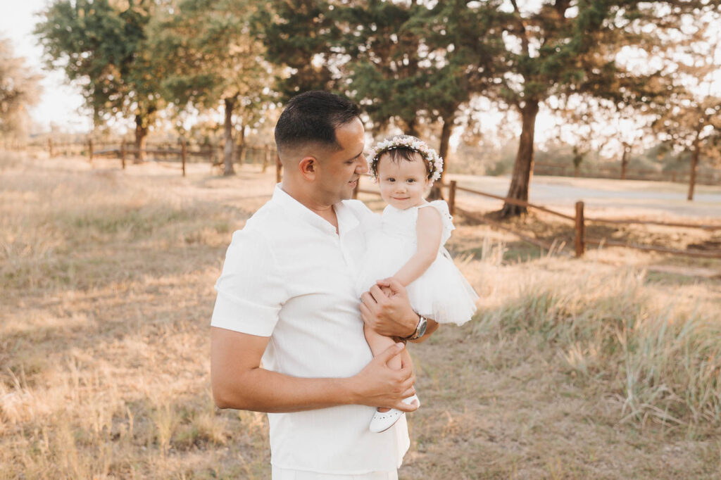 How I Create Natural Family Photos Without Awkward Posing 2 Father Holding His Smiling Toddler Daughter In A Golden Grassy Field With Warm Evening Sunlight And Trees In The Background.